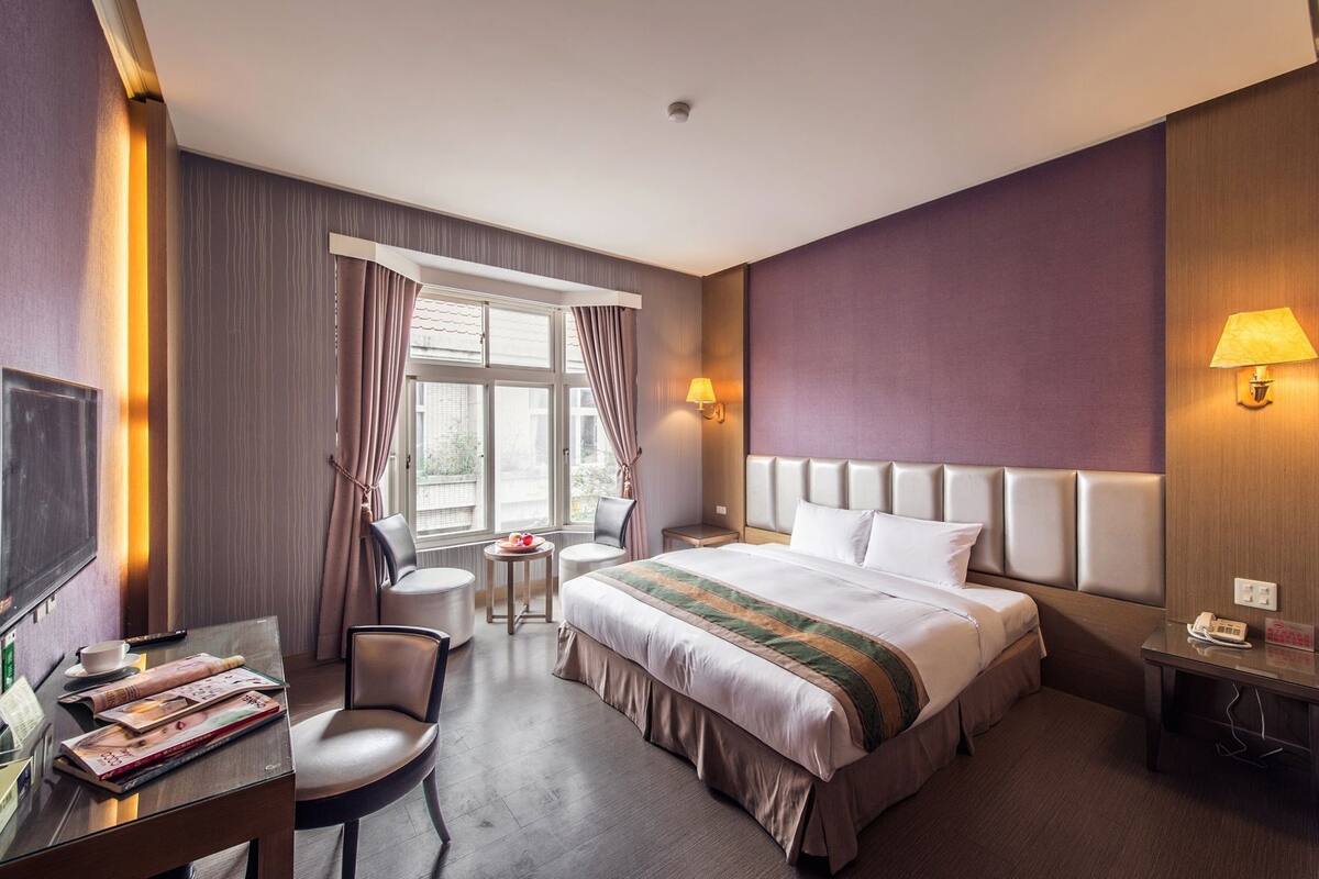 A hotel room with a bed, a desk with a chair, a small table, and a window with curtains.