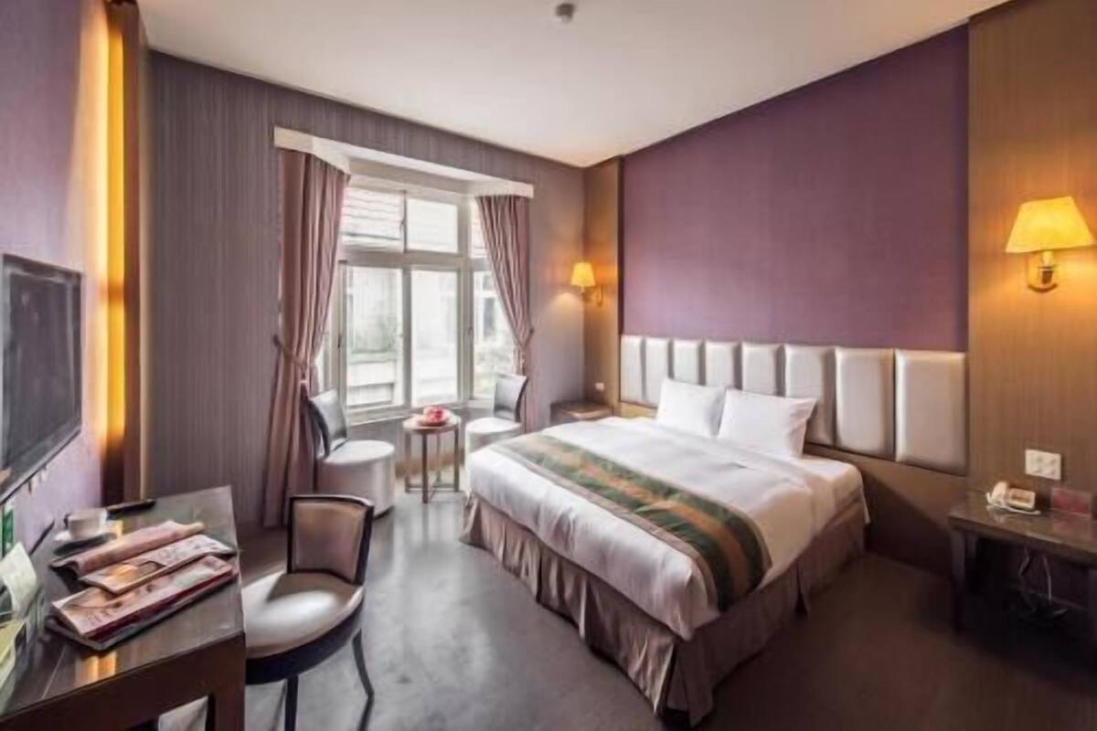A hotel room with a bed, a desk with a chair, a small table, and a window with curtains.