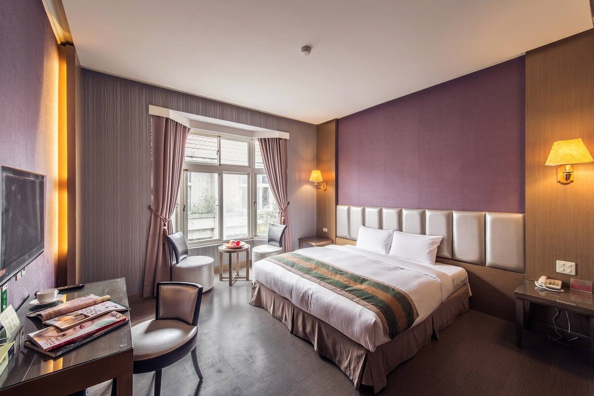 A hotel room with a bed, a desk with a chair, a small table with a chair, a window with curtains, and a wall-mounted lamp.