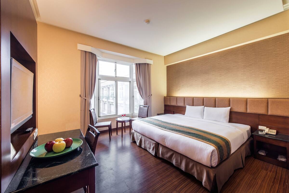 A hotel room with a large bed, a desk with a plate of fruit, a chair, a small table, and a window with curtains.