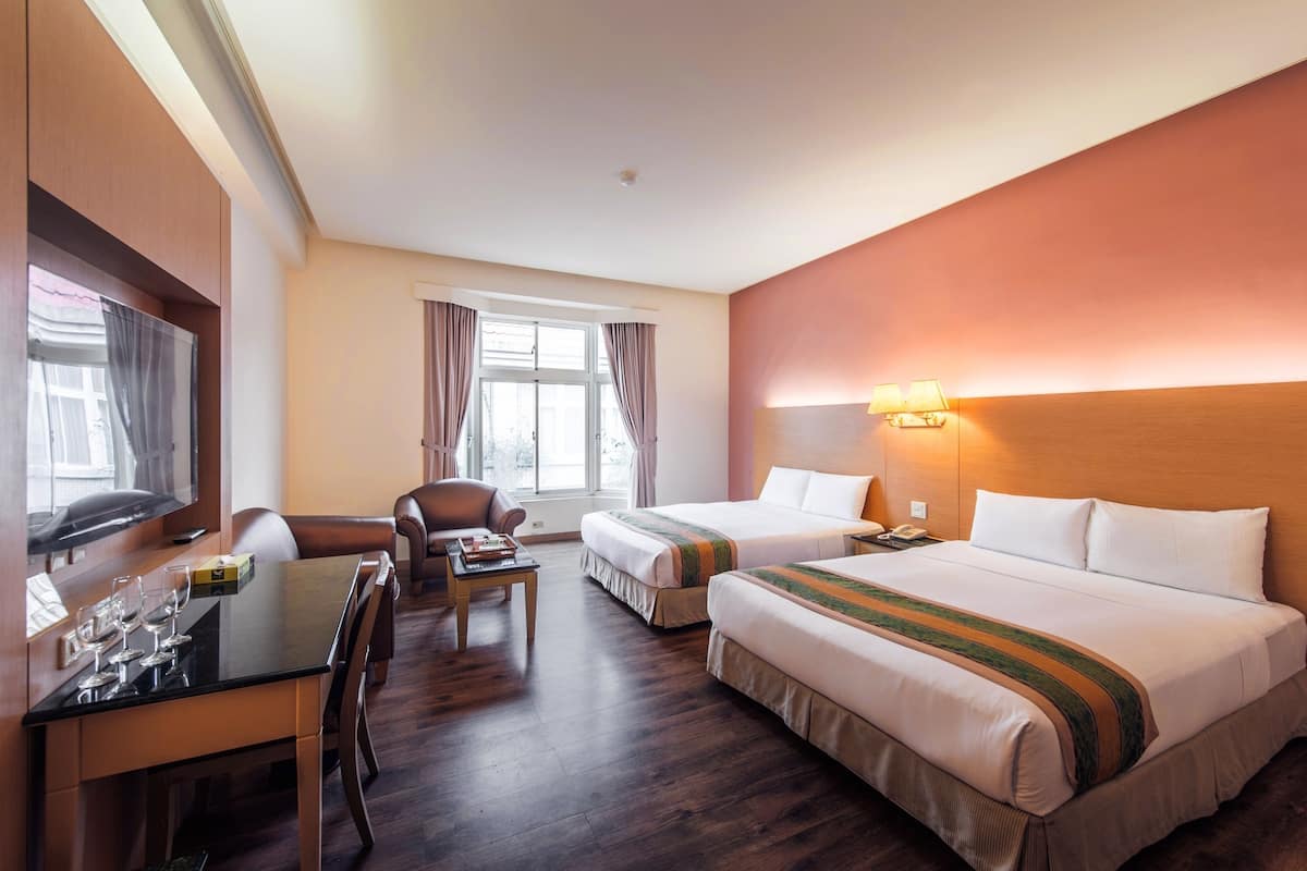 A hotel room with two beds, a desk with a chair, a TV, and a window with curtains.