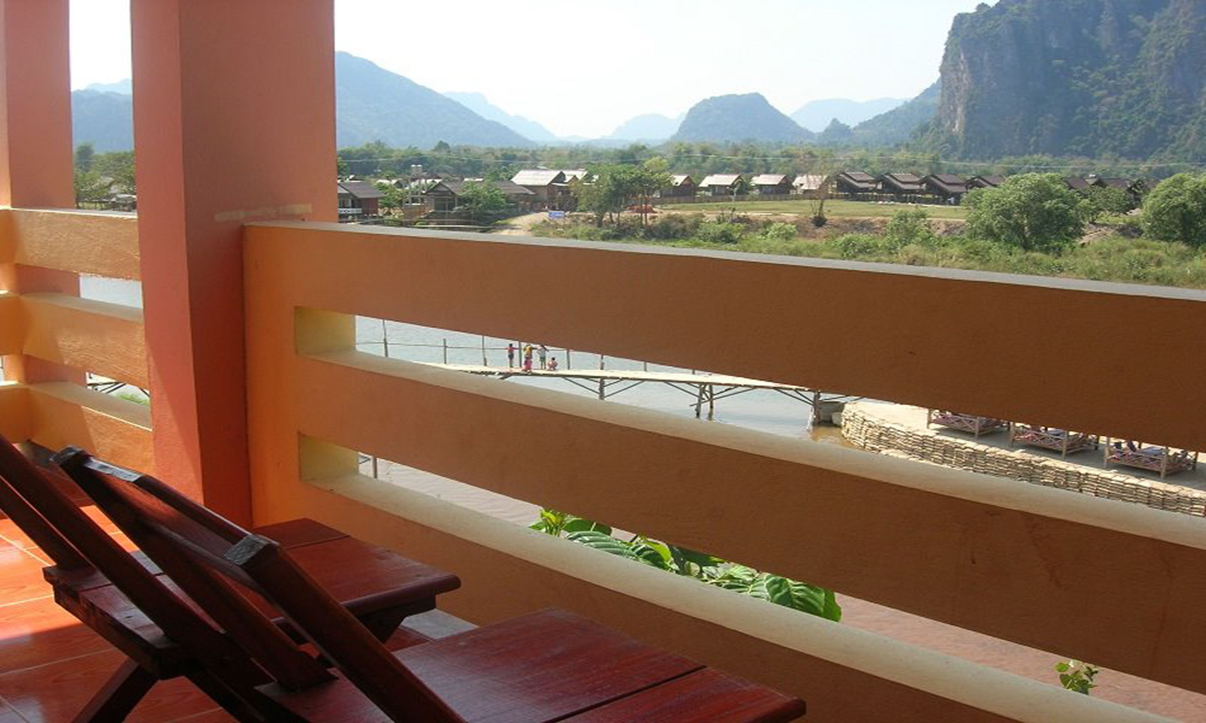 A balcony with a wooden bench, overlooking a river with traditional houses and mountains in the background.