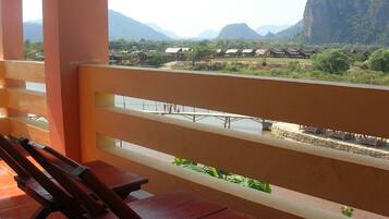 A balcony with a wooden bench, overlooking a river with traditional houses and mountains in the background.