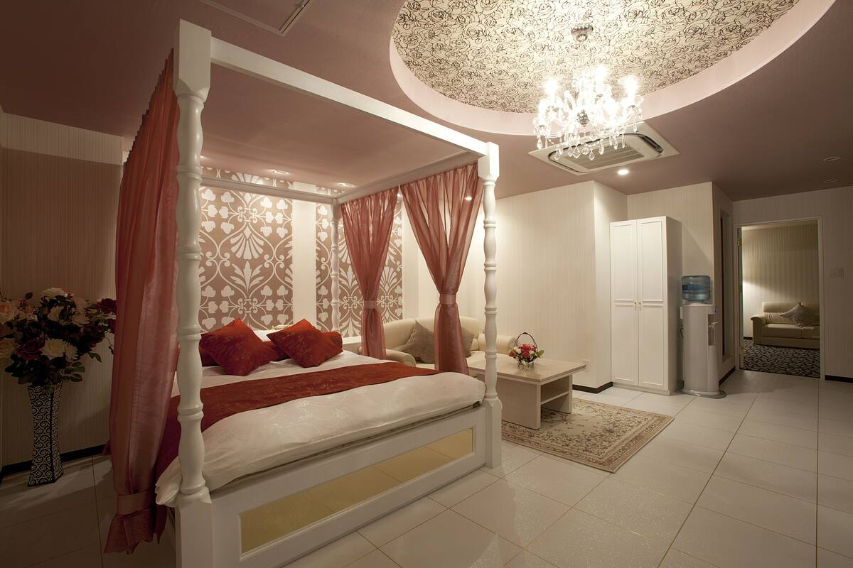 A four-poster bed with a canopy, a chandelier, and a water dispenser in a well-lit room with a patterned ceiling and a water feature.