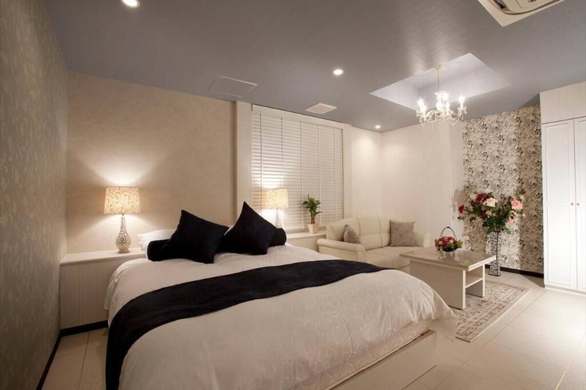 A modern bedroom with a large bed, bedside tables, a sofa, a chandelier, and a window with blinds.