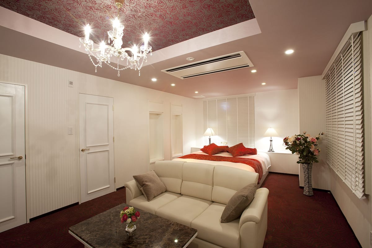 A hotel room with a chandelier, a bed with red pillows, a sofa, a table with a vase, and a window with blinds.