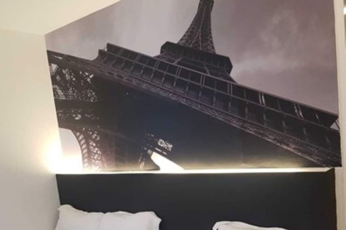 Quarto de hotel com duas camas de solteiro, uma pintura grande da Torre Eiffel e uma cabeceira moderna.