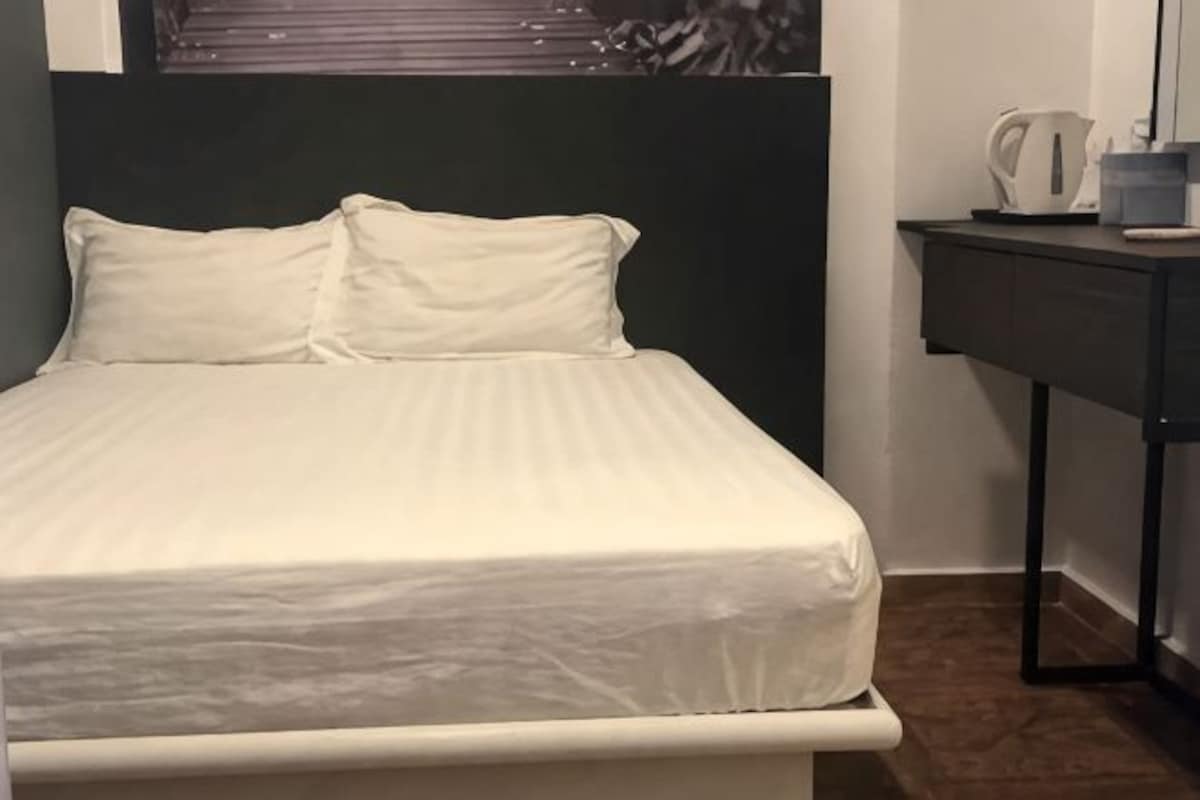 A bed with white bedding, a dark headboard, a small table with a kettle, and a mirror on a wall.