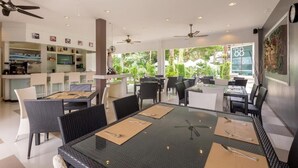 A modern dining area with glass tables, black and white chairs, ceiling fans, and a view of greenery outside. - The Regent Studio Kamala (Kamala)