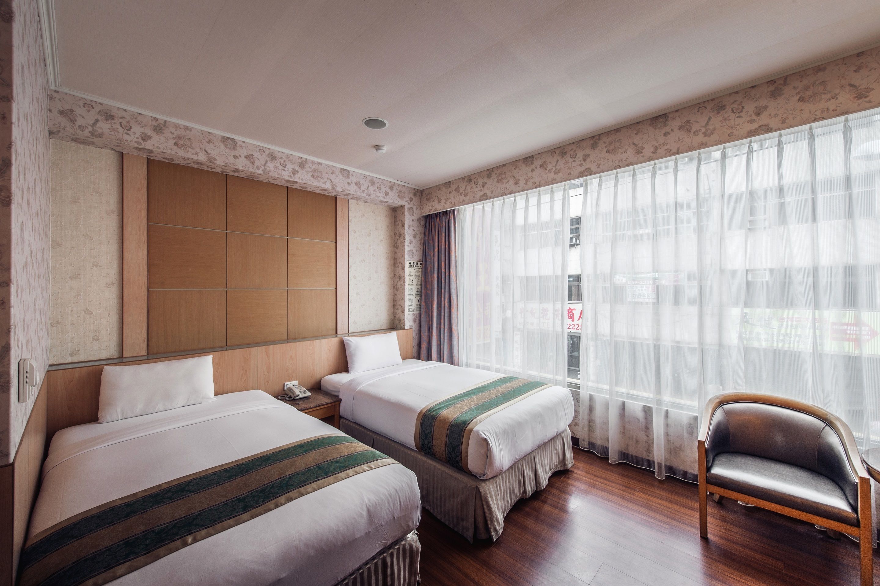 A hotel room with two beds, a wooden headboard, a chair, and a large window with sheer curtains.