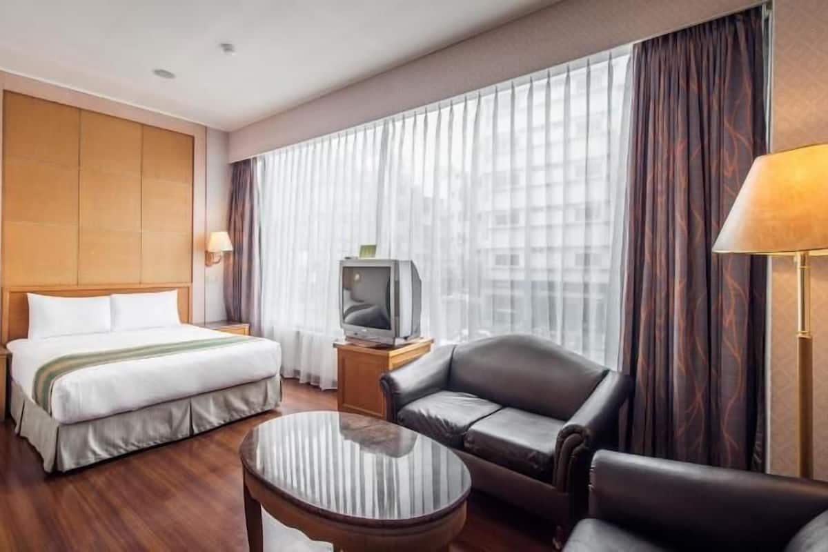 A hotel room with a bed, a TV on a wooden side table, a leather armchair, a round coffee table, and a large window with curtains.