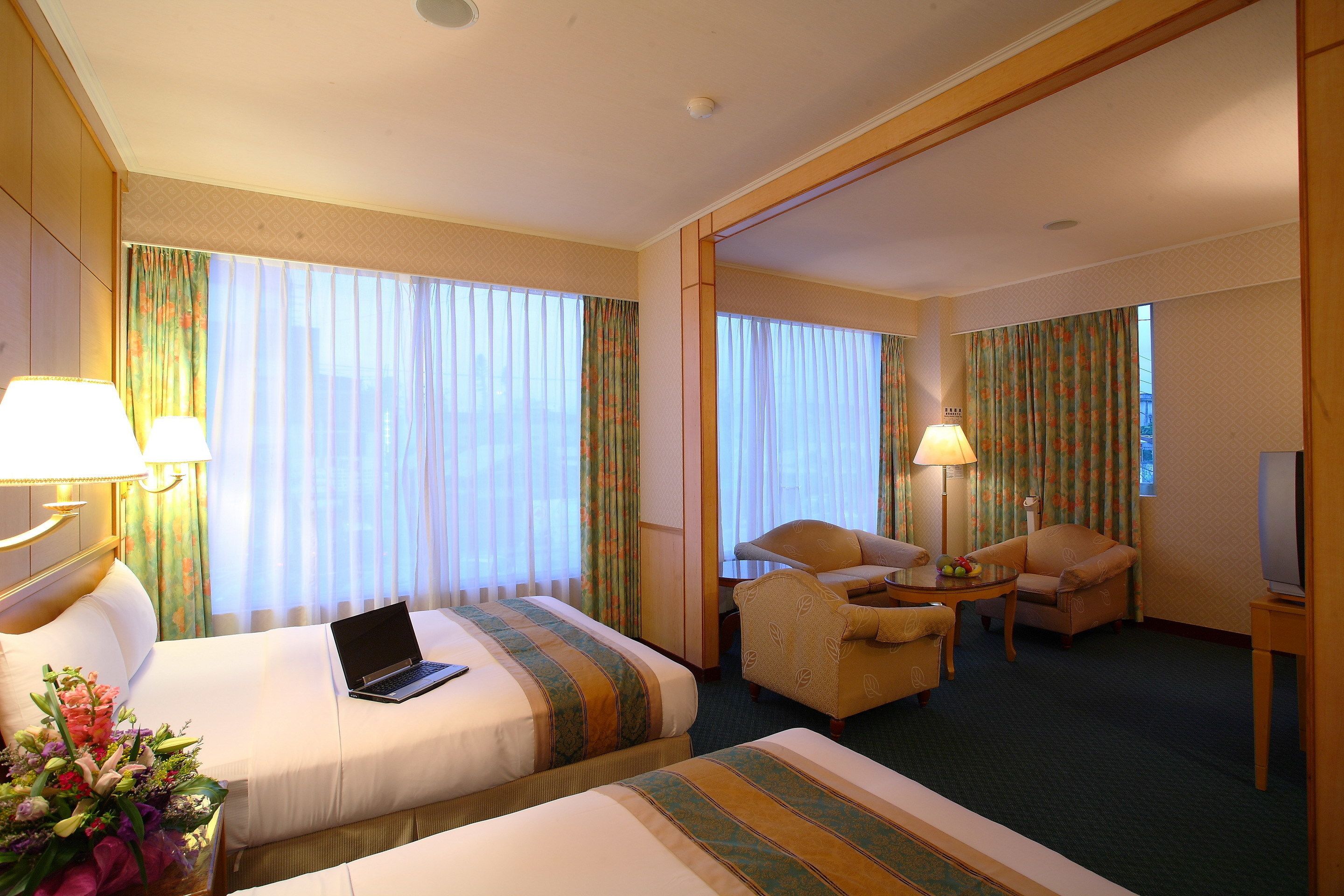 A hotel room with two beds, a desk, a chair, a TV, and a lamp.