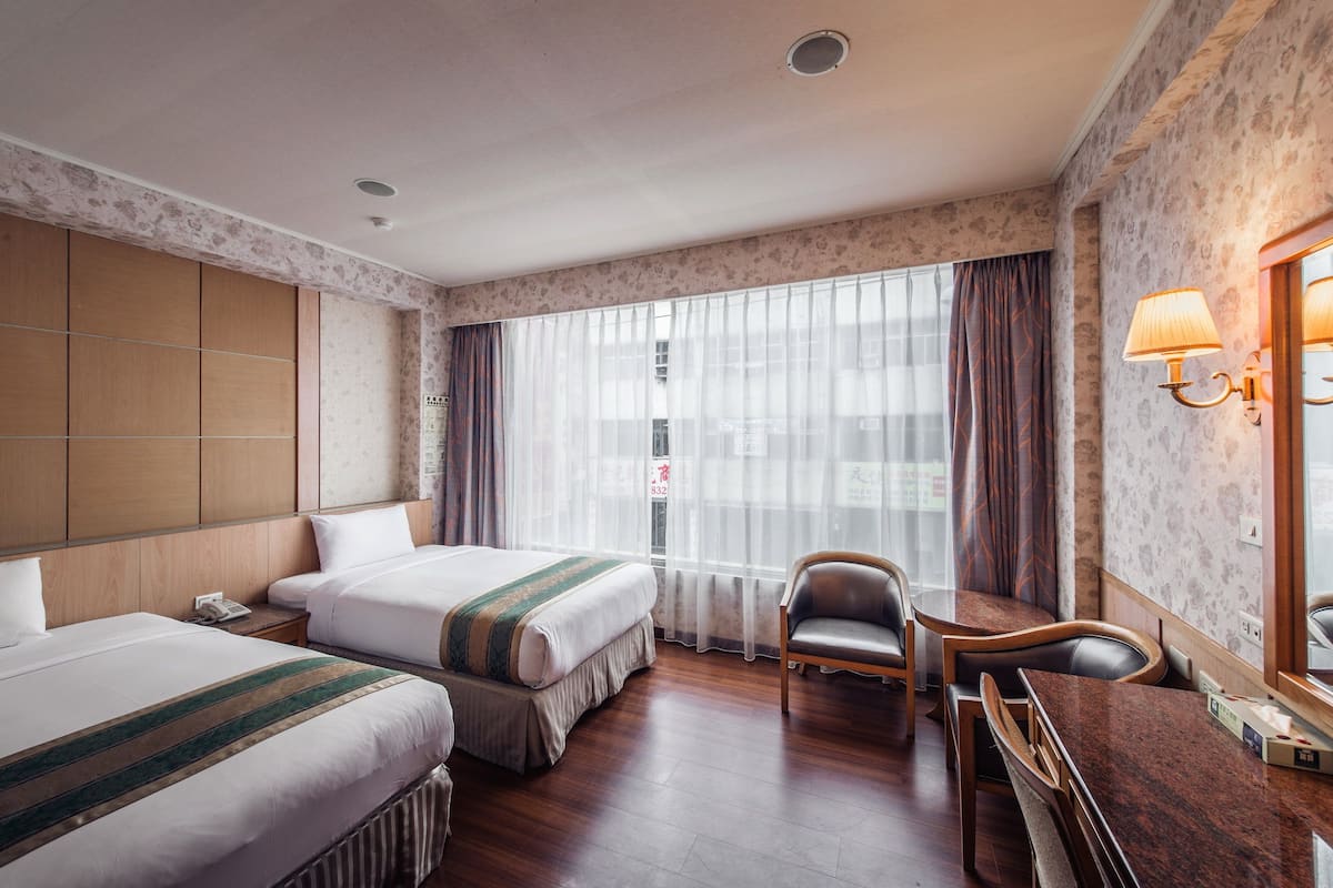 A hotel room with two beds, a wooden desk, a chair, and a large window with curtains.