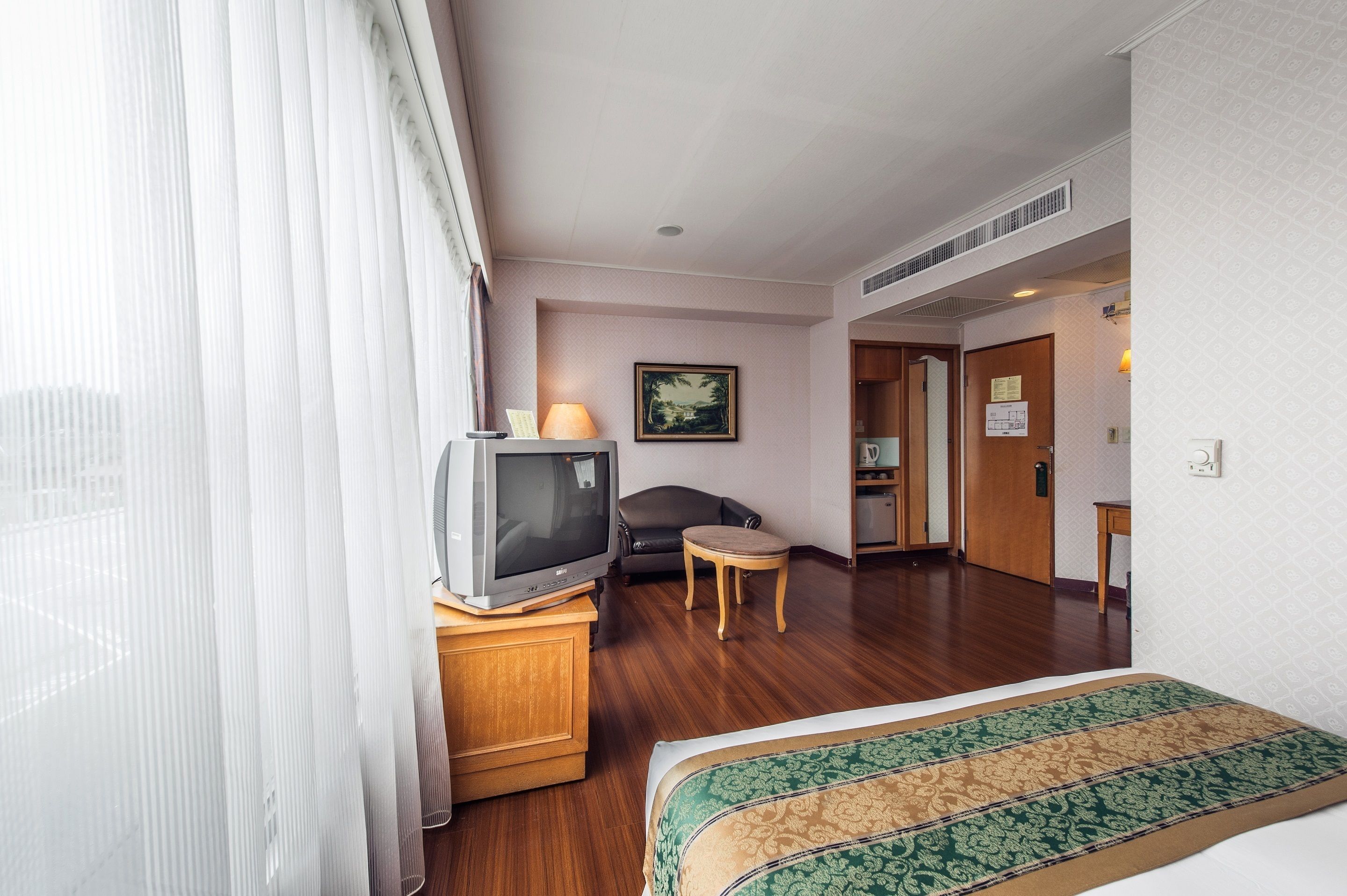 A hotel room with a bed, a television on a wooden cabinet, a small table, a chair, and a door leading to another room.