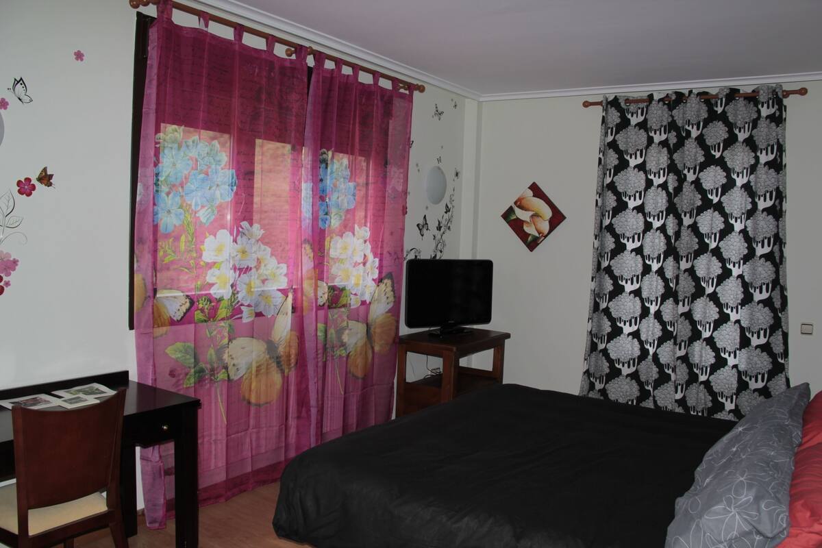 A bedroom with a bed, a television on a wooden stand, a desk with a chair, and a pink floral curtain.