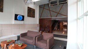 A modern lobby with a grey sofa, wooden tables, and a TV screen. - Ramedo Hotel (Makassar)