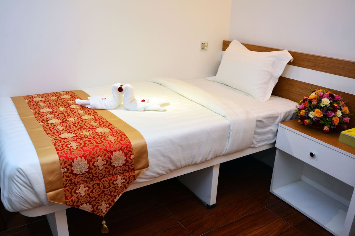 A neatly made bed with a red and gold patterned bedspread, a white pillow, and a bouquet of flowers on the nightstand.