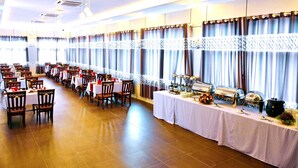 Un salón de banquetes con varias mesas dispuestas para comer, una barra de buffet con diversas opciones de comida y un amplio ventanal con cortinas.
