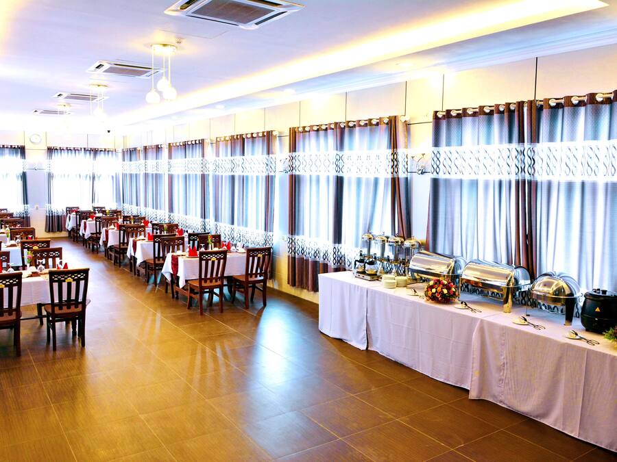 A banquet hall with multiple tables set for dining, a buffet line with various food items, and a large window with curtains.