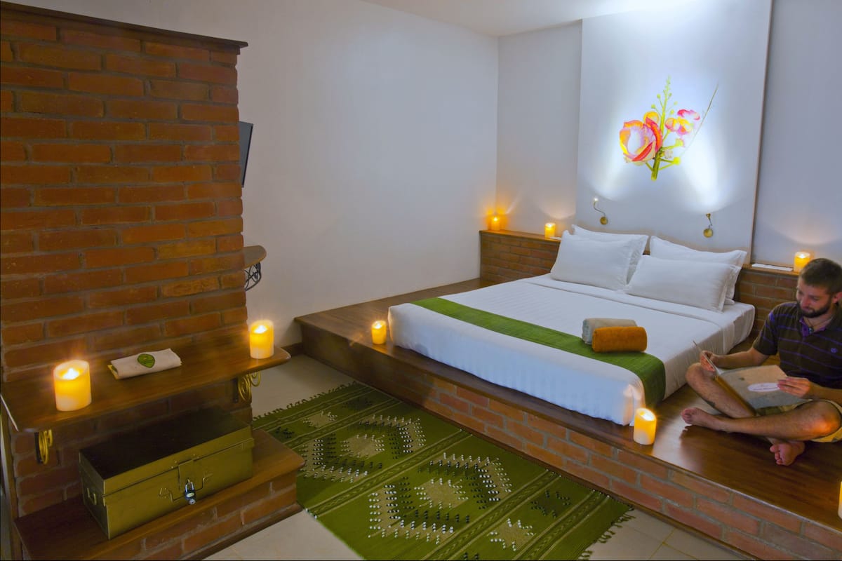 A modern hotel room with a brick fireplace, a bed with white linens, and a green throw blanket. There are candles on the floor and a wall-mounted artwork of flowers.