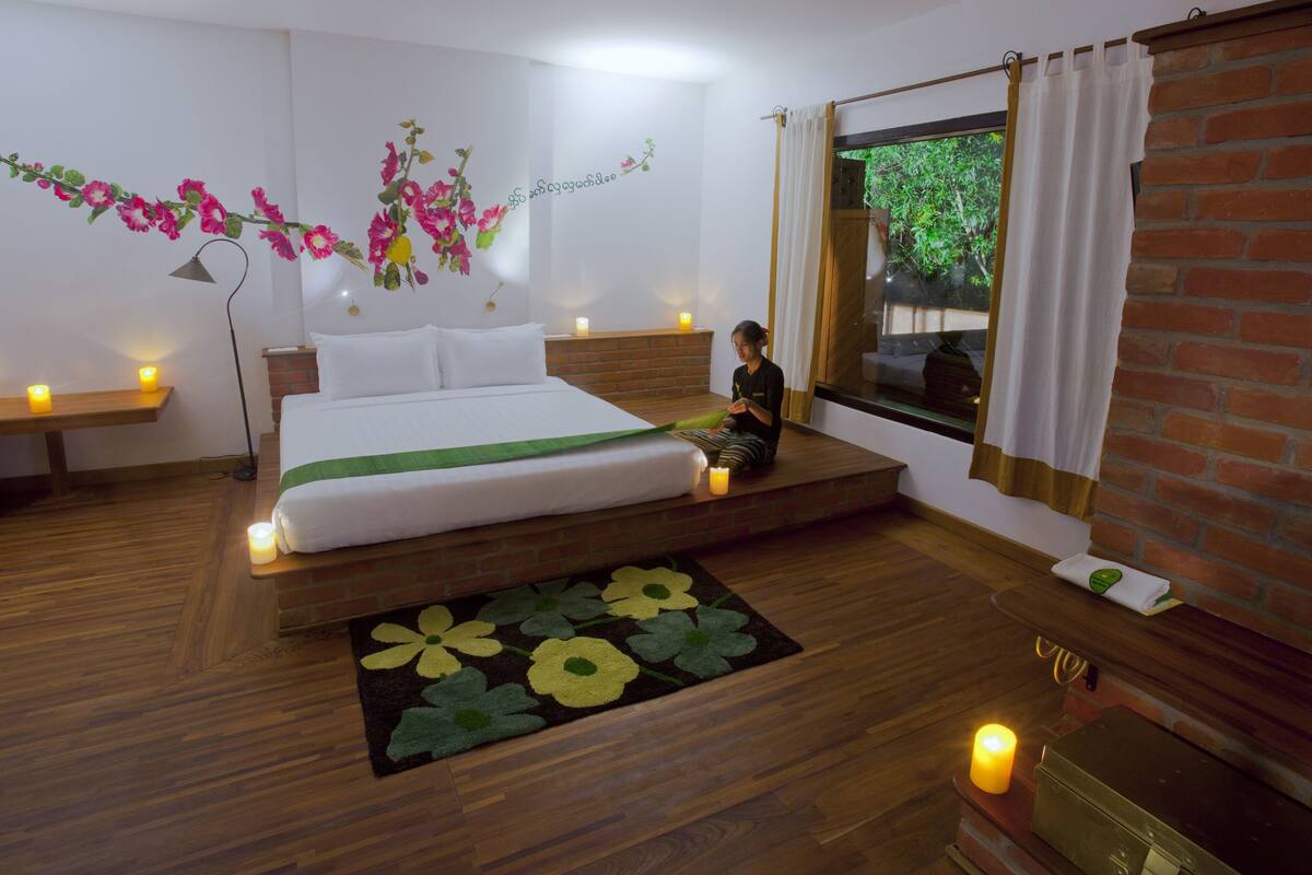 A bedroom with a bed, a nightstand, a window with curtains, a wooden floor, and a wall with floral decorations.