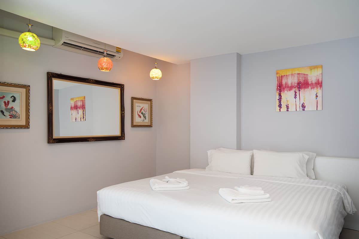 A hotel room with a large bed, white linens, a framed artwork on the wall, and a ceiling air conditioner.