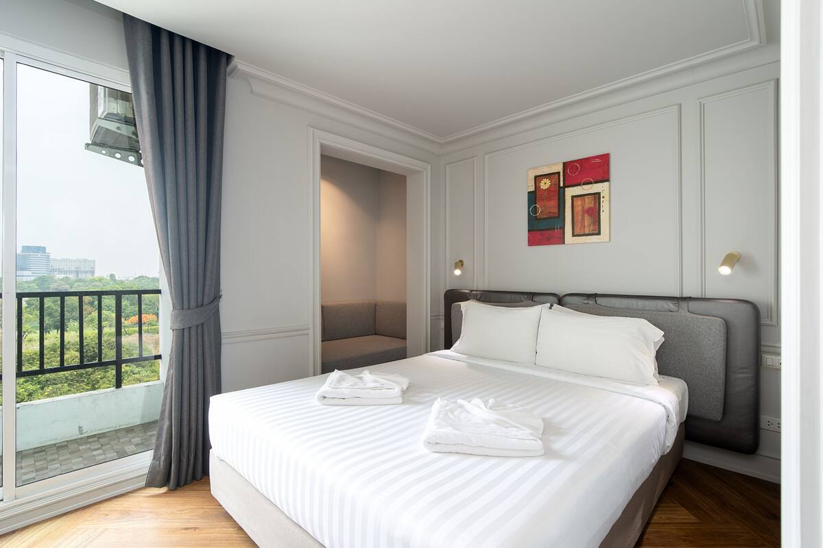 A hotel room with a large bed, a view of greenery through a balcony, and a painting on the wall.