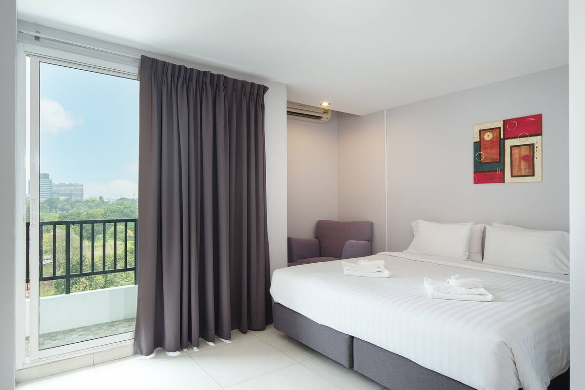 A hotel room with a large bed, a chair, a balcony with curtains, and a city view.