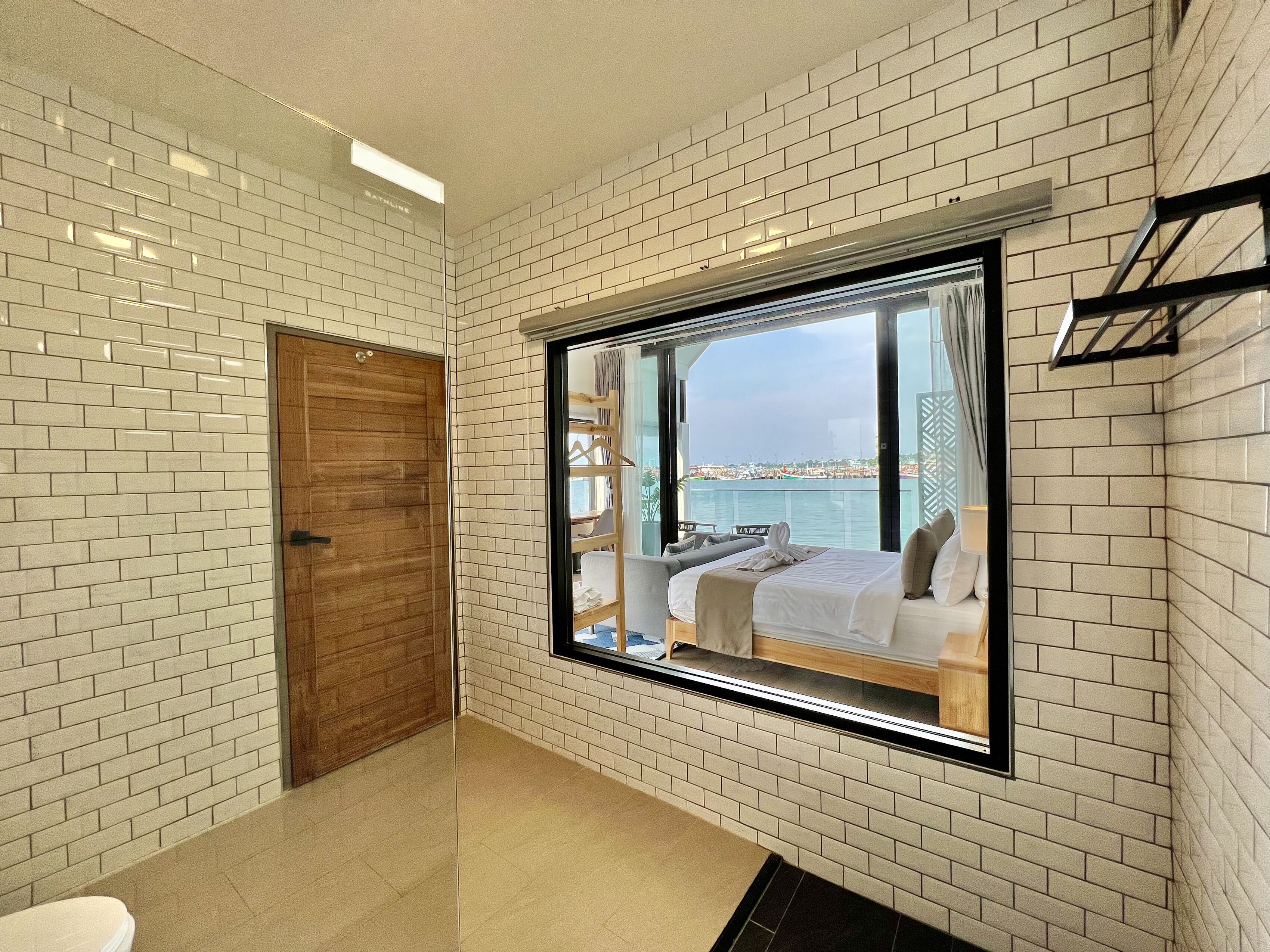 Un baño con paredes de azulejos blancos, una puerta de madera y un ventanal que ofrece vistas a un balcón con una cama y un paisaje urbano.