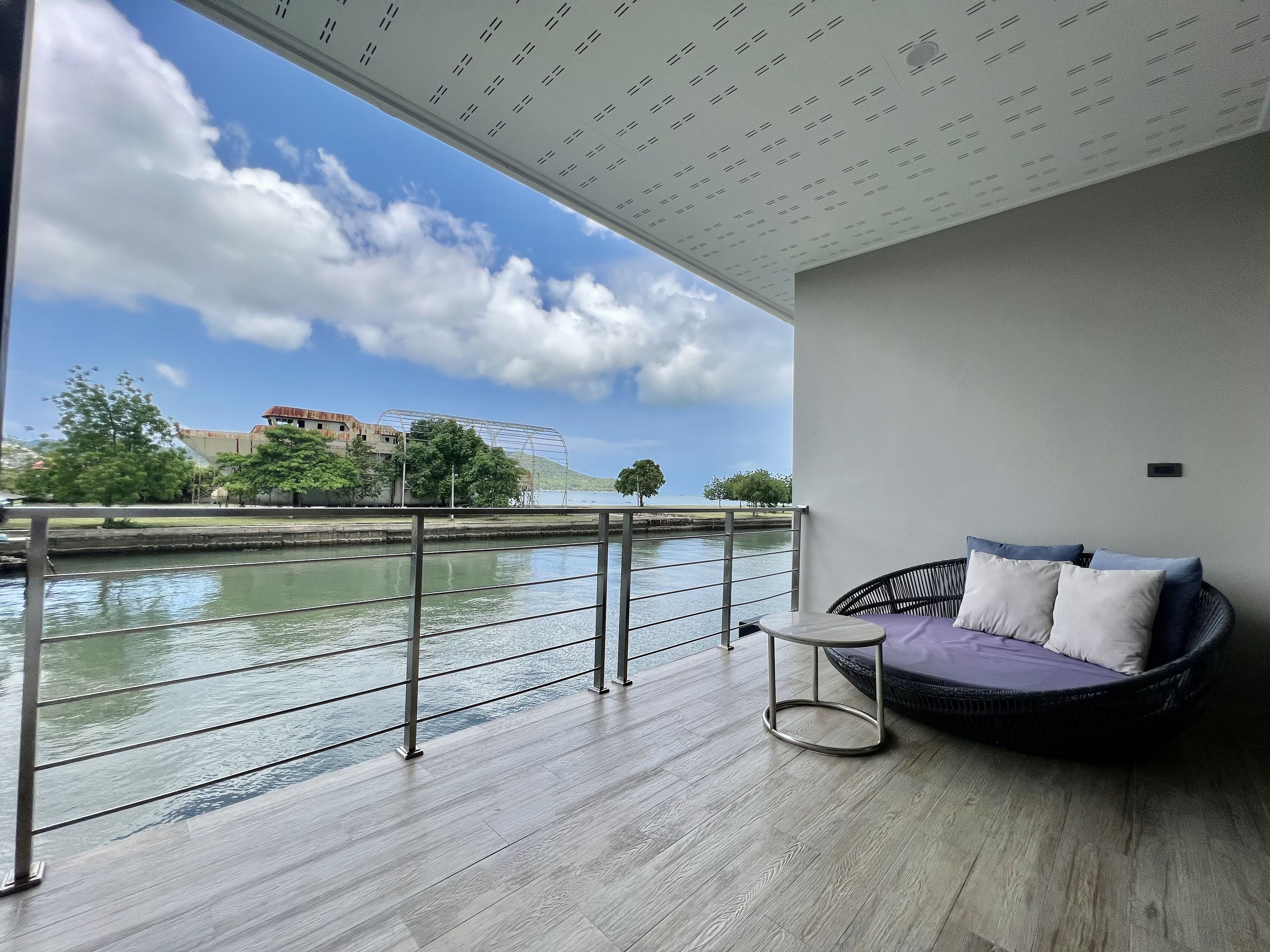 Un balcón con vistas a un río, barandilla de metal y un sofá de mimbre con cojines.