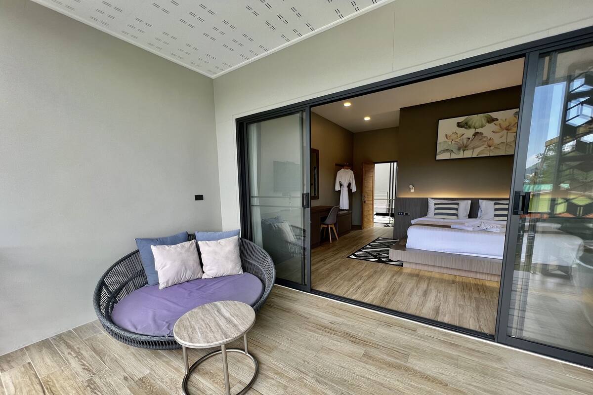 Een moderne kamer met een glazen deur naar een balkon, een ronde rieten zithoek met kussens, een klein rond tafeltje en een bed met een gestreept hoofdeinde.
