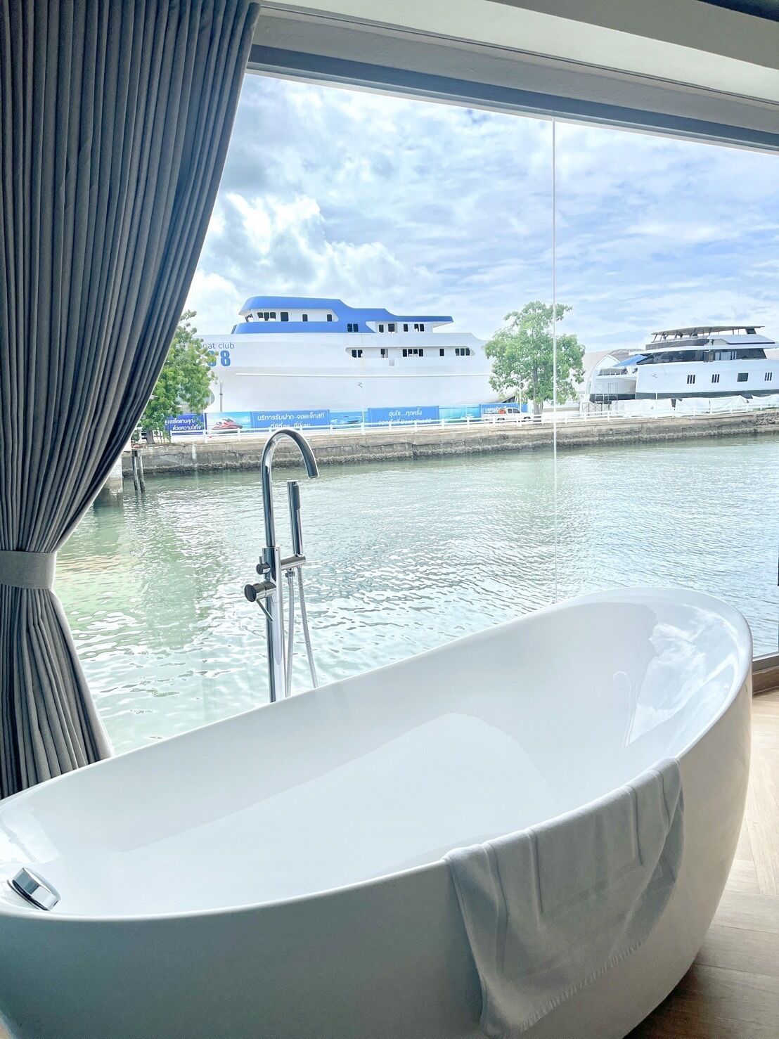 A modern bathroom with a freestanding bathtub, a window with curtains, and a view of a river with boats.