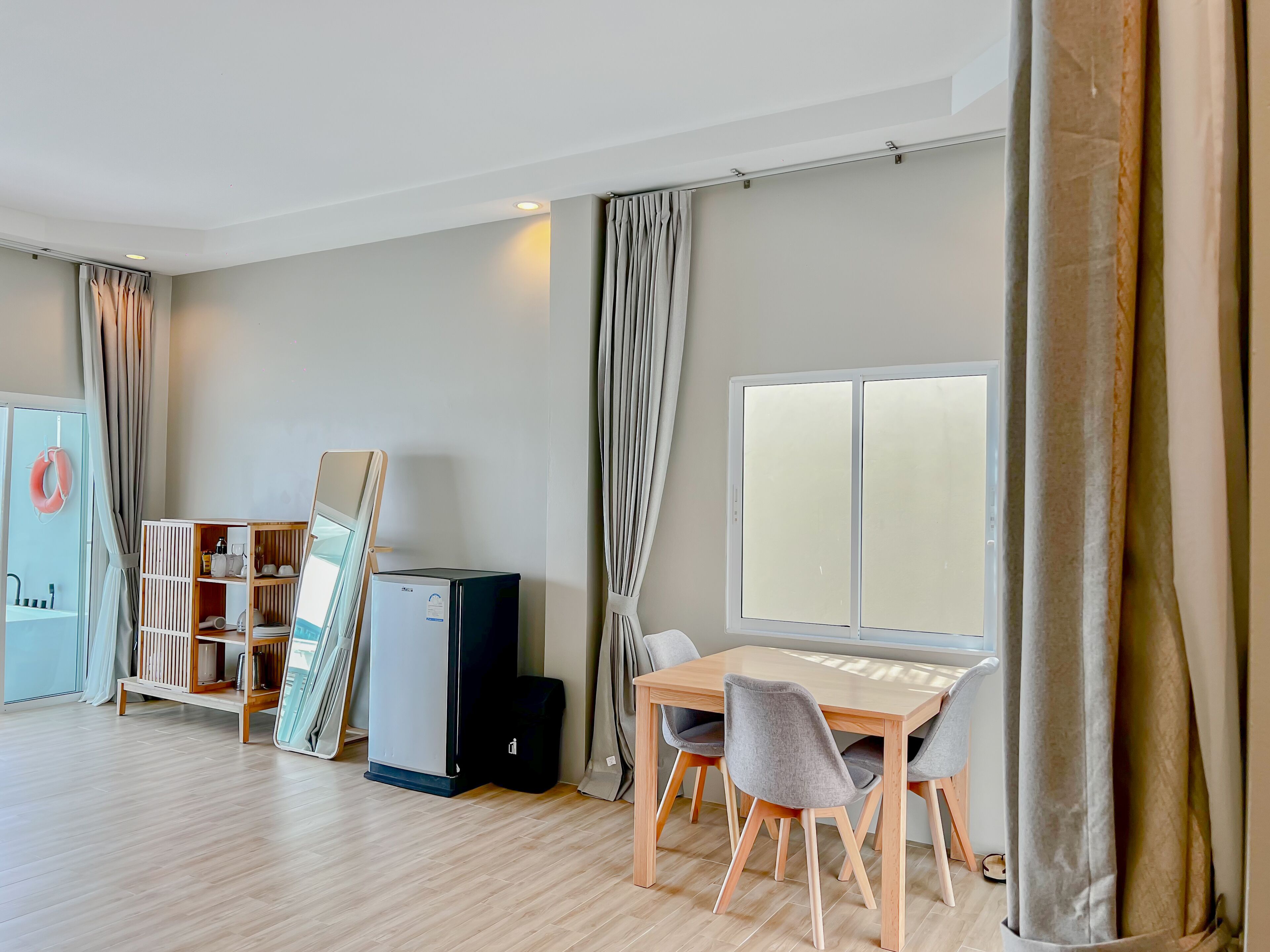 Une chambre d’hôtel avec un plancher en bois, une table à manger avec des chaises, un petit réfrigérateur, une armoire avec un miroir et une fenêtre avec des rideaux.
