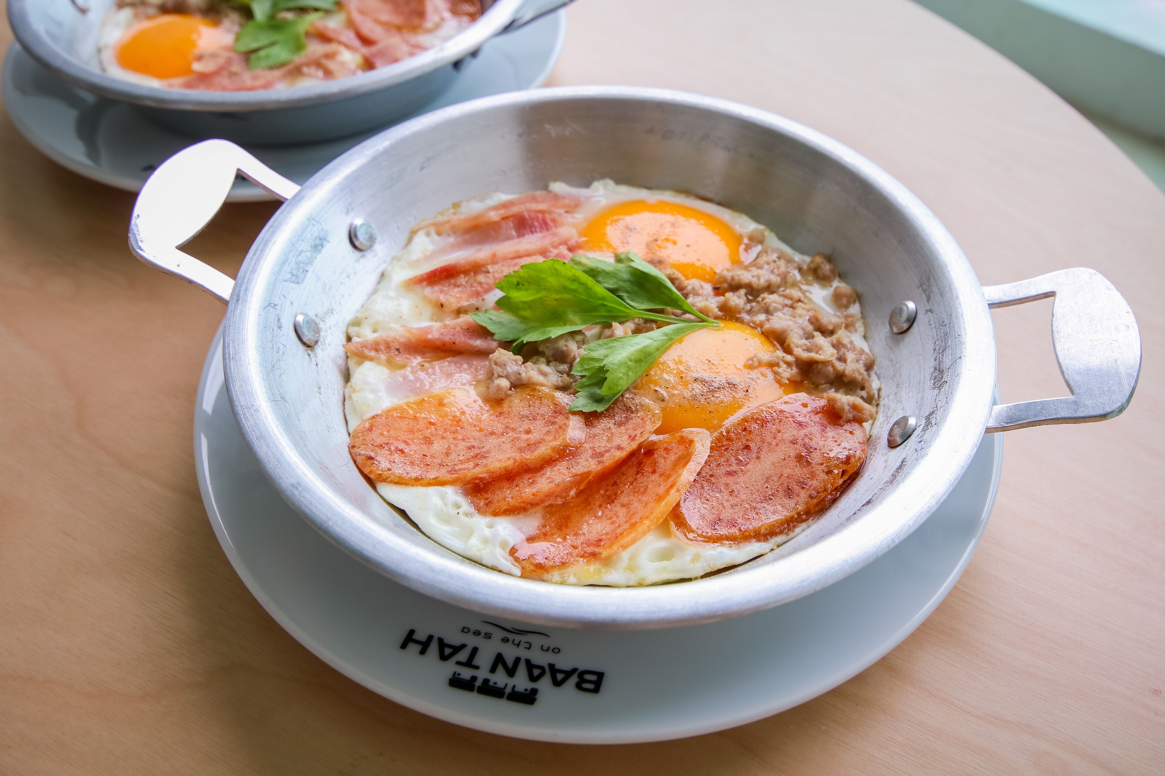 A metal bowl with a handle, containing a breakfast dish with eggs, meat, and a leaf garnish.