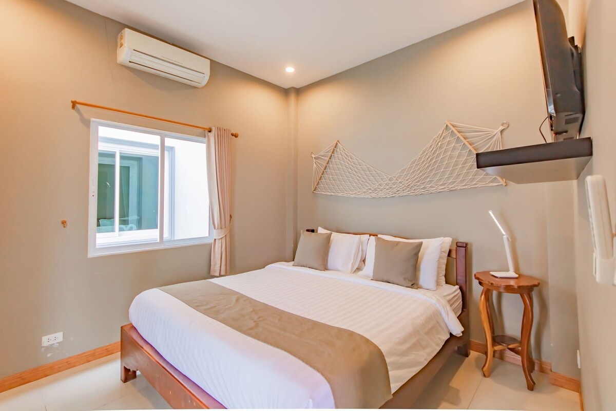 Une chambre à coucher comprenant un lit, une fenêtre, un climatiseur, une table de chevet et un téléviseur fixé au mur.