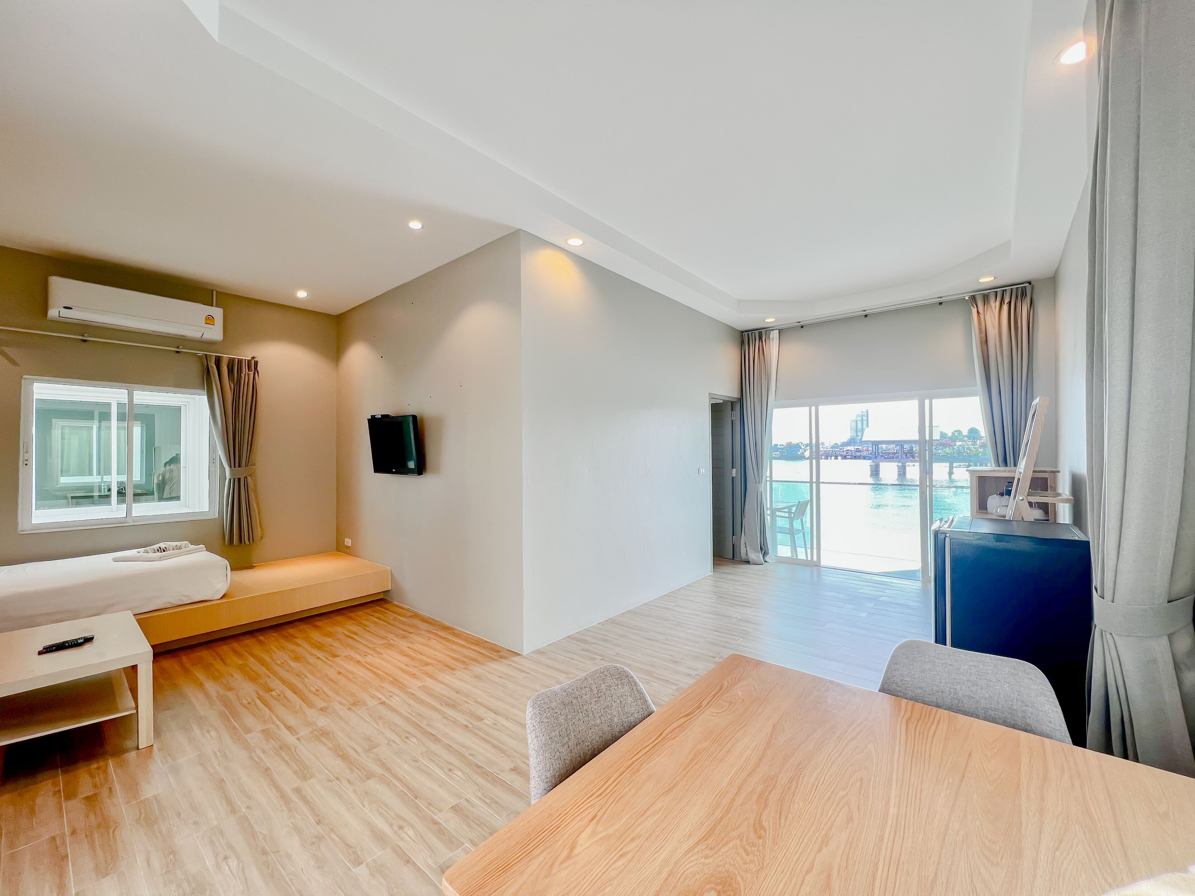 Habitación de hotel moderna con piso de madera, cama, escritorio, silla, televisión y un amplio ventanal con vista a un cuerpo de agua.