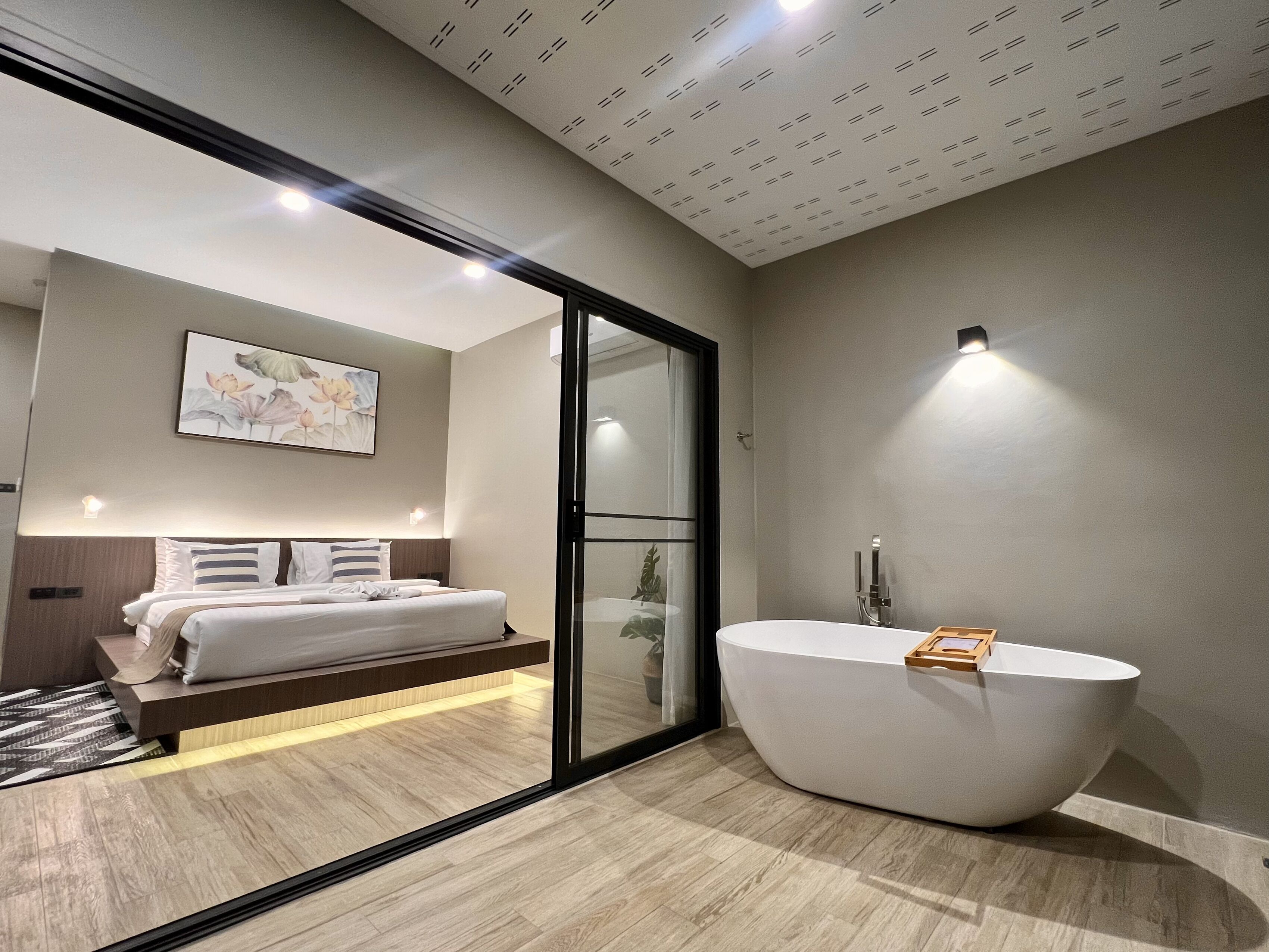 A modern hotel room with a large bed, a glass door leading to a balcony, a large freestanding bathtub, and a wall-mounted light fixture.