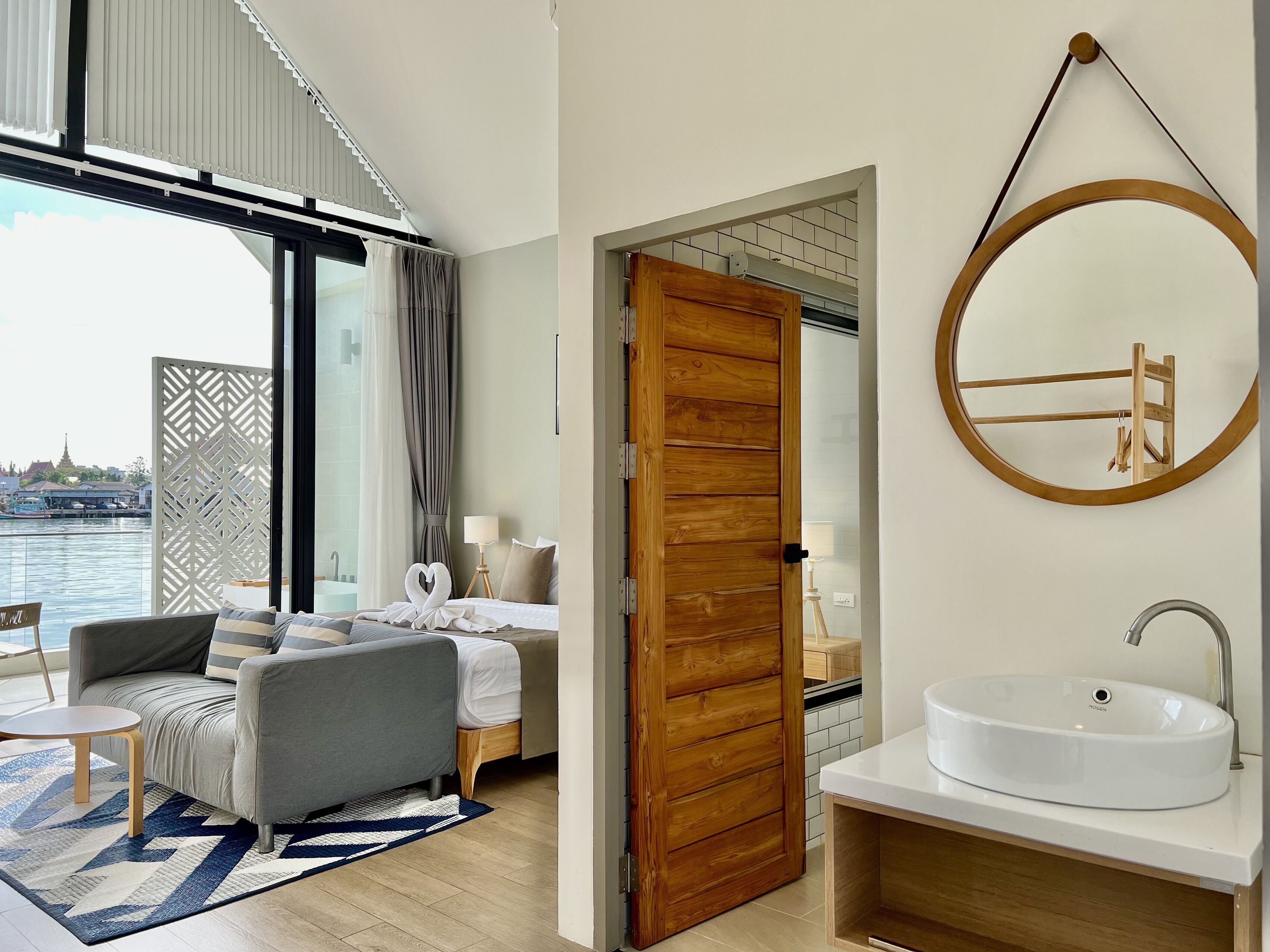 A modern hotel room with a large window, a round mirror, a wooden door, and a white sink.