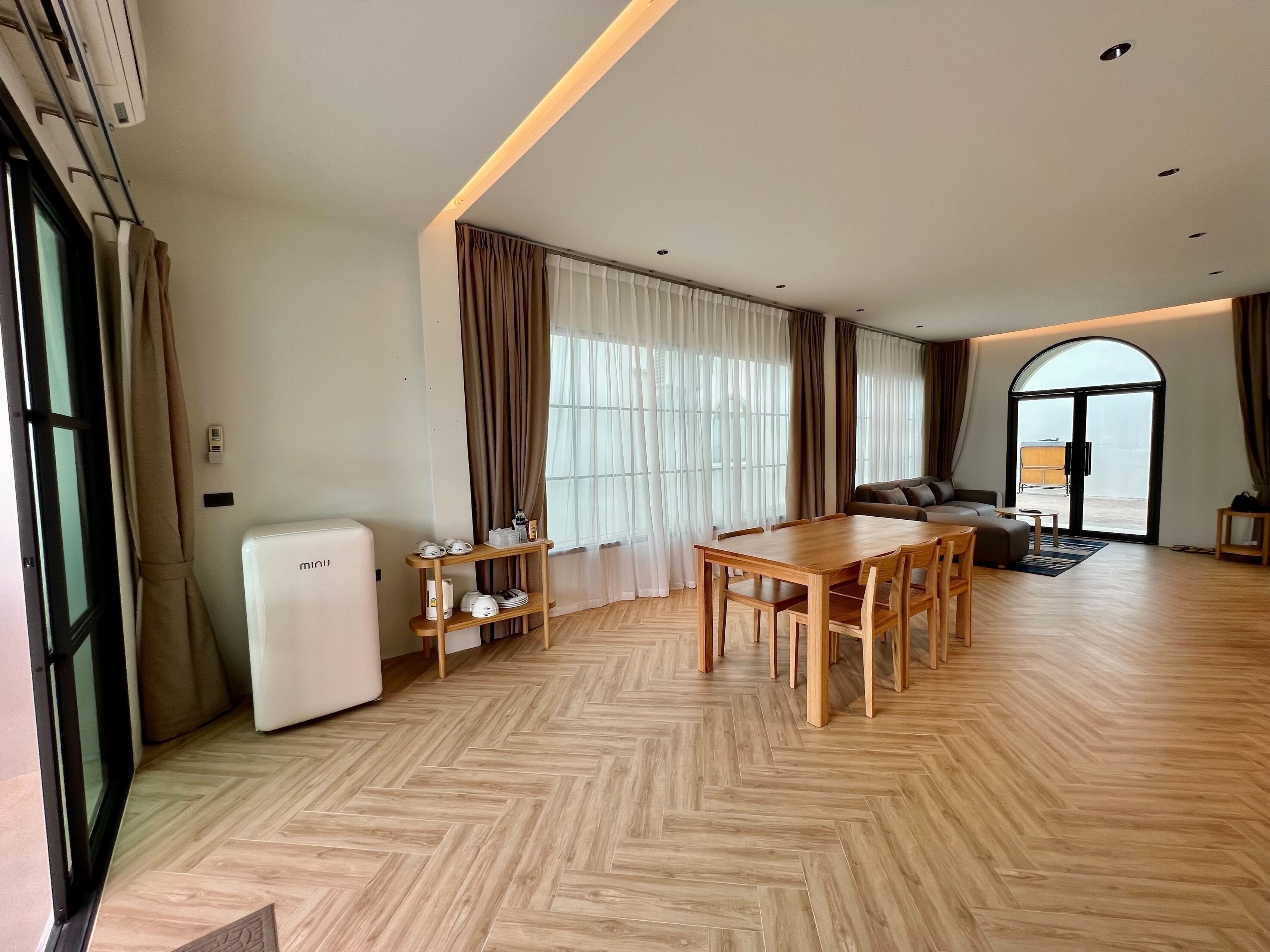 A spacious living area with a wooden herringbone floor, a dining table with chairs, a sofa, and a large window with curtains.