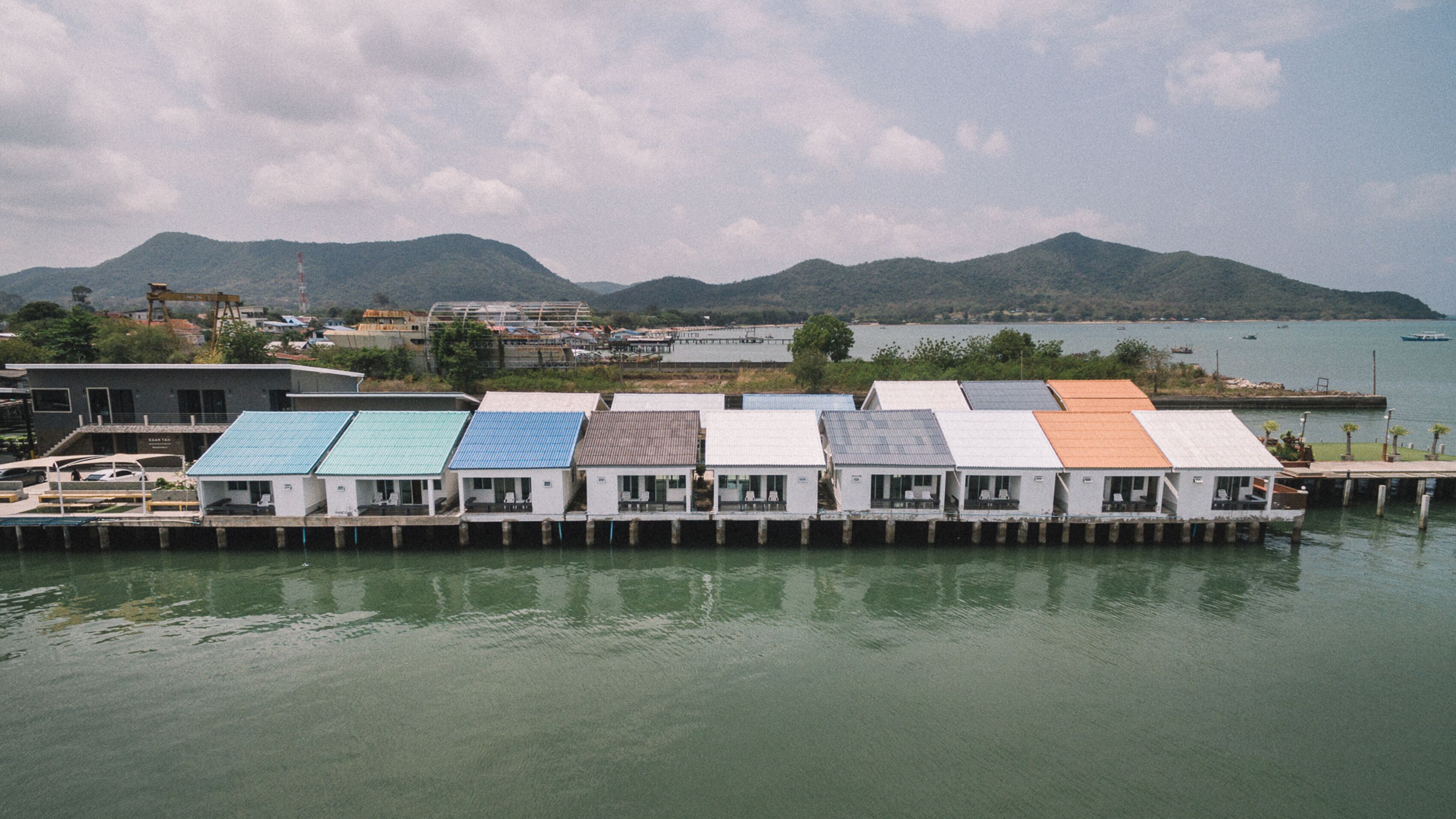 A row of small, single-story buildings with flat roofs, likely residential or commercial units, situated along a waterfront with a backdrop of hills and a body of water.