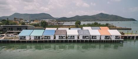 A row of small, single-story buildings with flat roofs, likely residential or commercial units, situated along a waterfront with a backdrop of hills and a body of water.