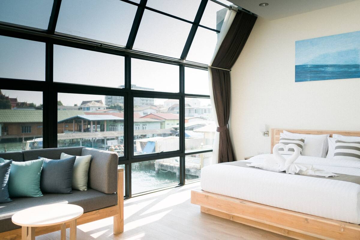 Een hotelkamer met een groot raam, een bed met witte lakens, een houten hoofdbord en uitzicht op gebouwen.