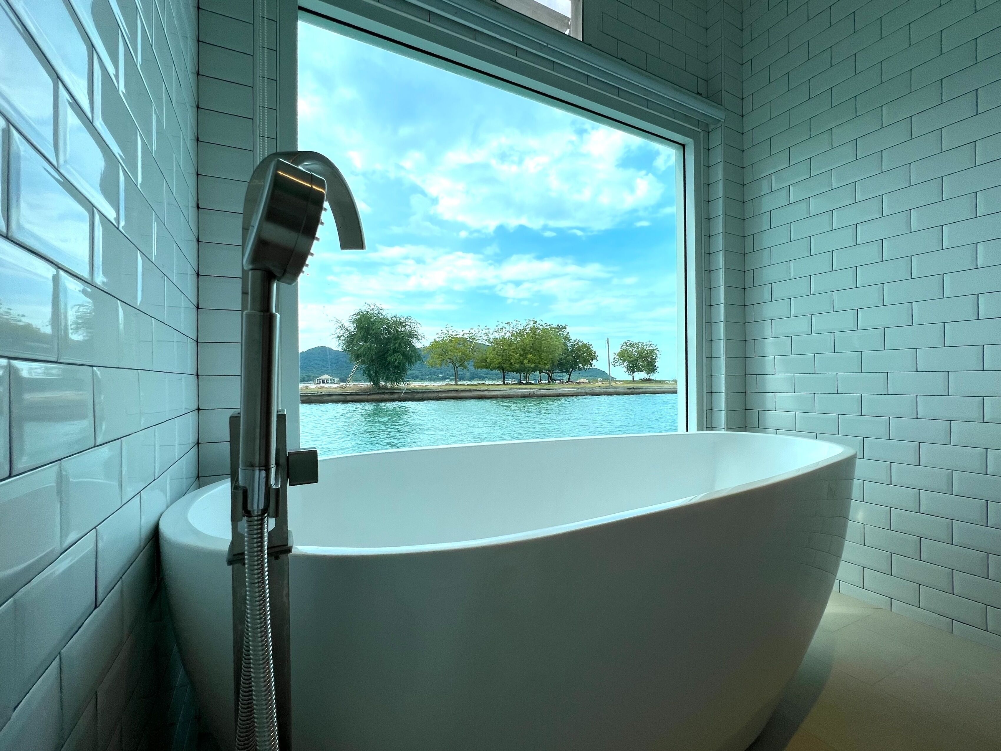 Un bagno moderno con una vasca da bagno ampia, una finestra vista acqua e alberi, e pareti rivestite di piastrelle bianche.
