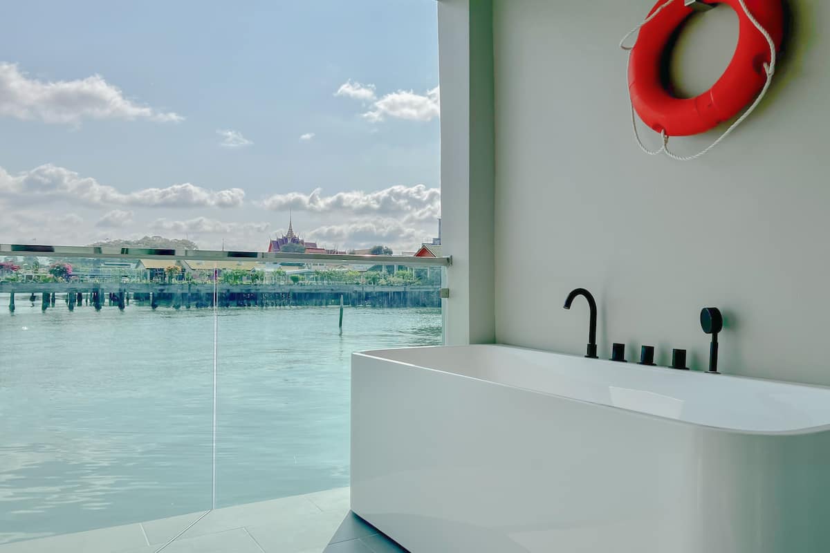 Een moderne badkamer met een groot bad, een rode reddingsboei aan de muur, en uitzicht op een waterpartij met gebouwen op de achtergrond.