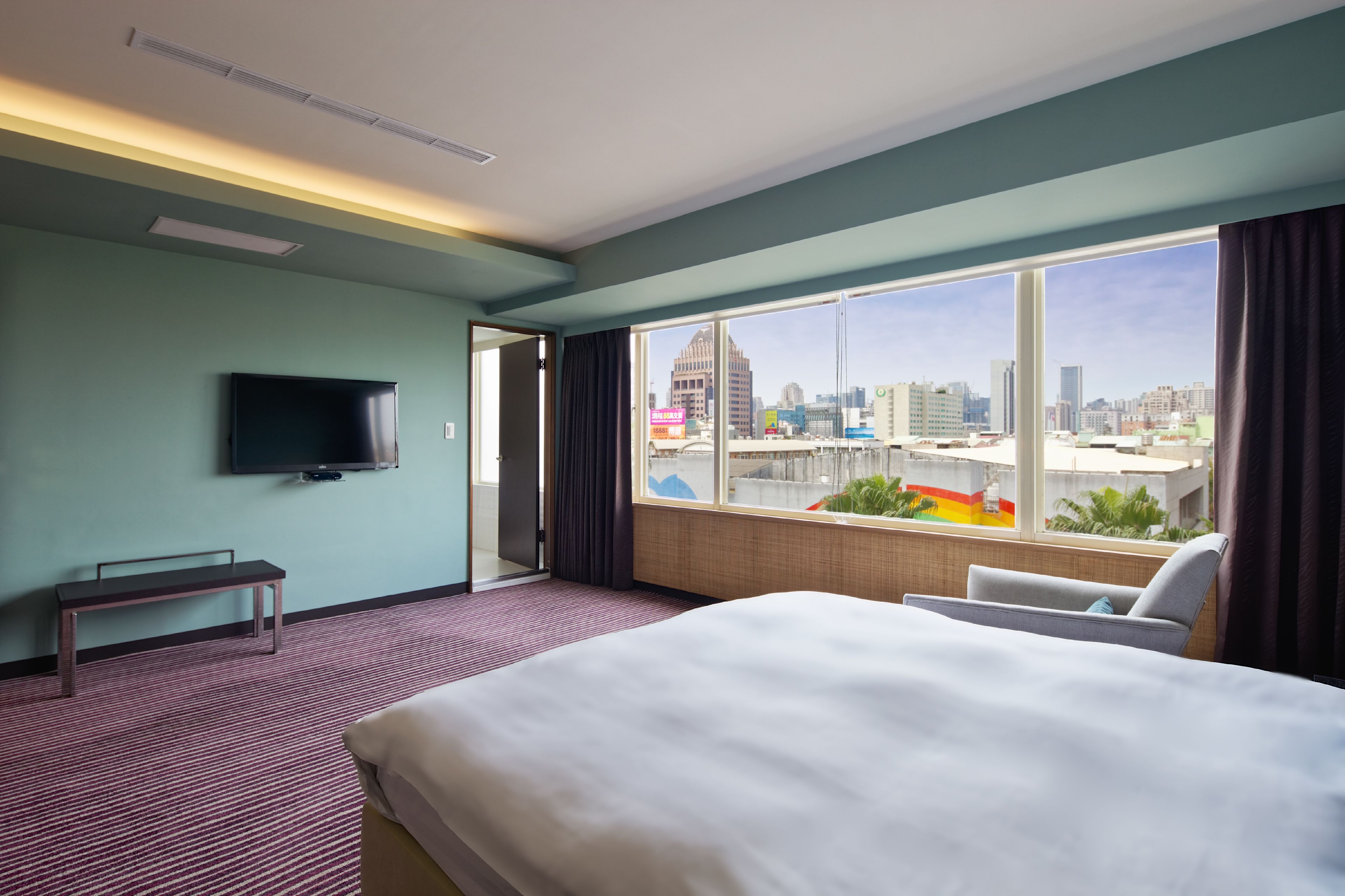 A hotel room with a bed, a bench, a TV, and a large window offering a city view.