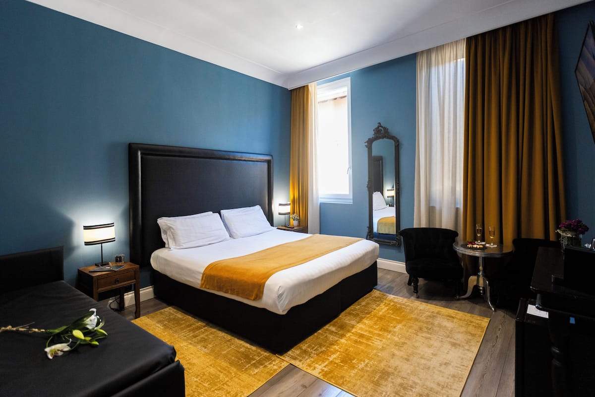 Habitación de hotel con una cama grande, una mesita de noche, un escritorio, una silla, un espejo y una ventana con cortinas.
