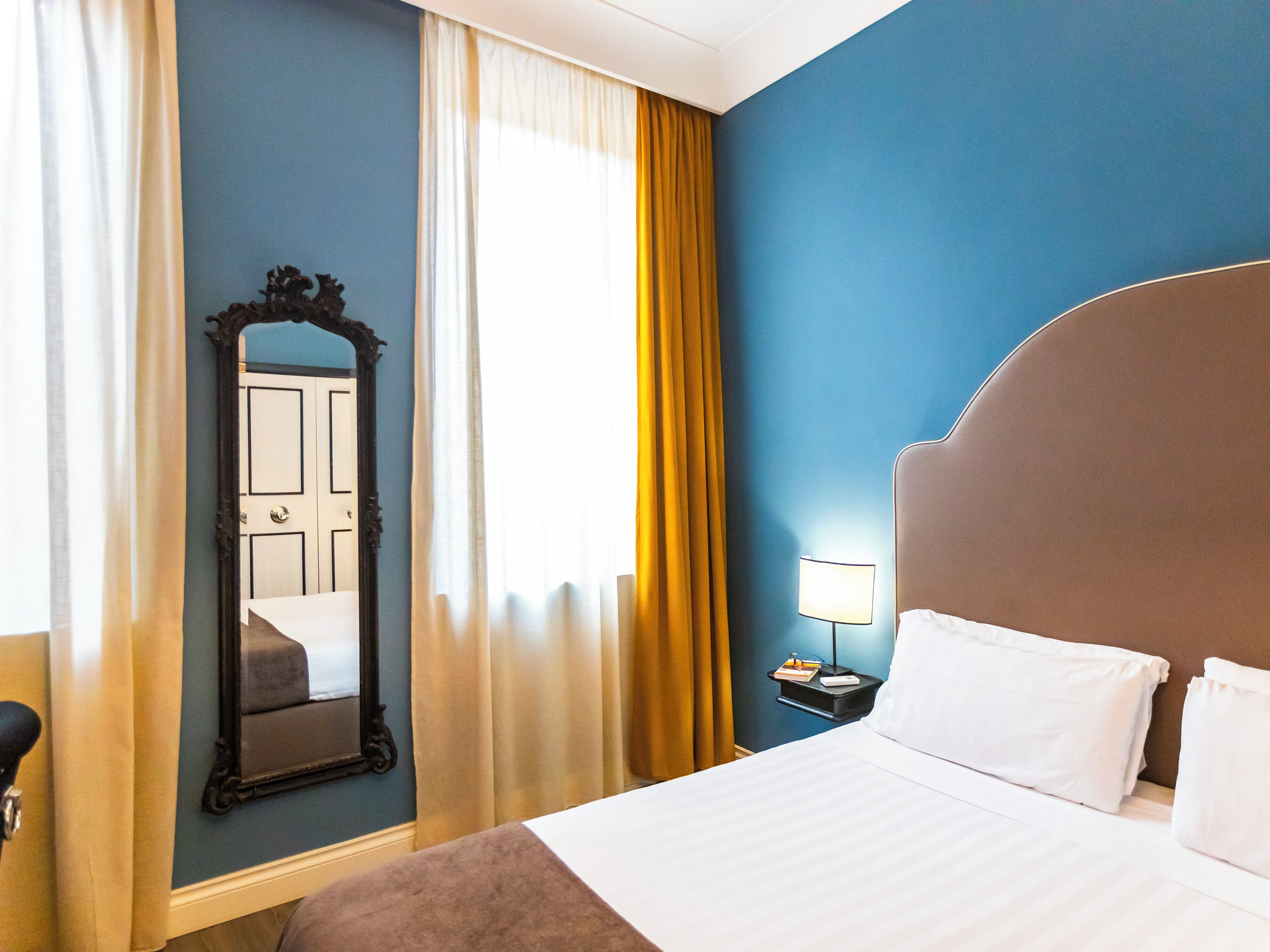 A hotel room with a large bed, a nightstand with a lamp, a mirror, and a door.