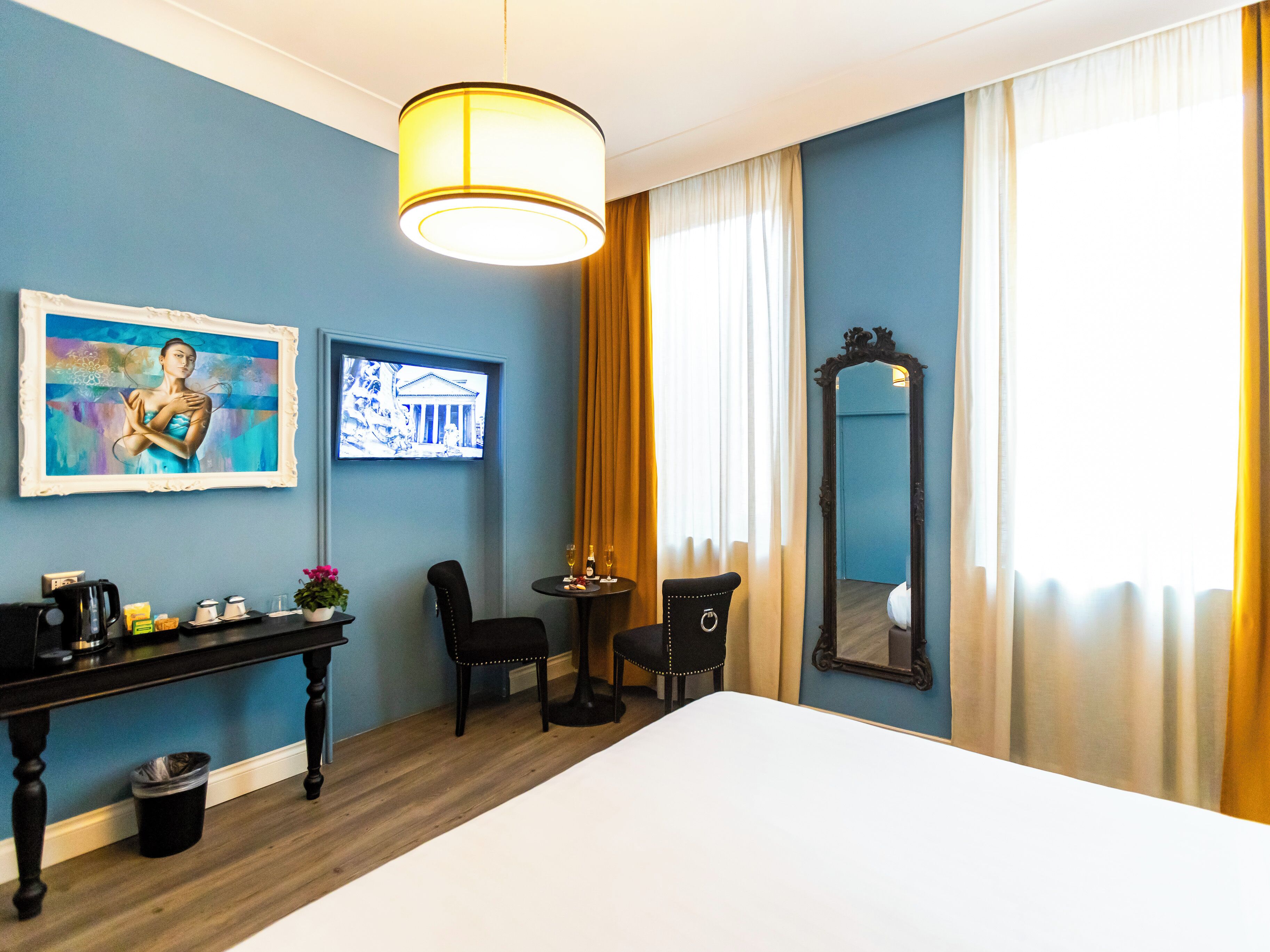 A hotel room with a bed, a desk with a coffee maker, a chair, a TV, a mirror, and a window with curtains.