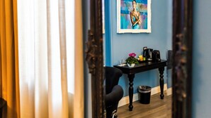 Una habitación con pared azul, un cuadro enmarcado, una silla negra, una mesita con cafetera y un cubo de basura.