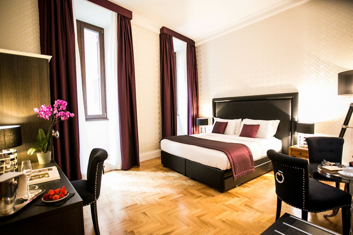 A hotel room with a large bed, a desk with a chair, a small table, and a potted plant.